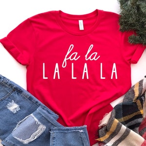 May include: Red t-shirt with white text that says "fa la LA LA LA" on a white background. The shirt is styled with blue jeans and a plaid scarf.
