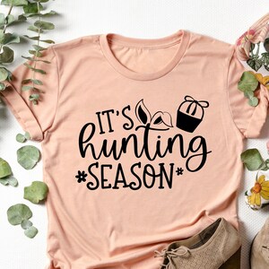 K&ouml;nnte beinhalten: Ein pfirsichfarbenes T-Shirt mit schwarzem Text, der "It's hunting season" lautet, mit einer kleinen Grafik eines Jagd-Hutes und einer Jagd-Weste.