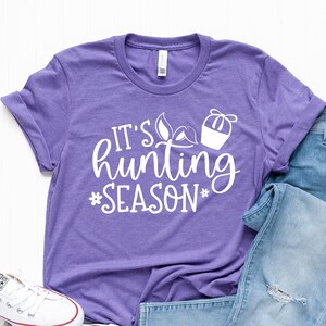 K&ouml;nnte beinhalten: Lilafarbenes T-Shirt mit wei&szlig;em Text, der "It's hunting season" lautet, mit einer kleinen Grafik eines Jagdkorbs und Bl&auml;ttern.