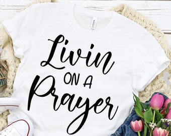 Peut inclure: T-shirt blanc avec un texte noir qui dit "Livin on a Prayer". Le t-shirt est posé à plat sur une surface en bois avec une paire de baskets blanches, une tasse à café, un jean et un bouquet de fleurs roses.