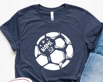 Può includere: Una maglietta blu navy con una grafica di un pallone da calcio bianco. Il pallone da calcio ha il testo "NAME 32" al suo interno.
