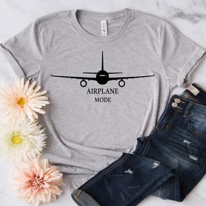 May include: A gray t-shirt with a black airplane silhouette and the text "AIRPLANE MODE" printed on the front. The shirt is laid out on a white surface with a pair of blue jeans and flowers.