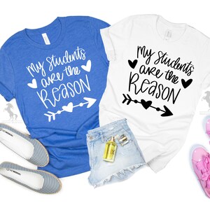 Op de afbeelding: Twee T-shirts met de tekst "Mijn leerlingen zijn de reden" erop. E&eacute;n shirt is blauw en de andere is wit. Beide shirts hebben een hart- en pijlontwerp.
