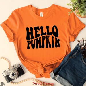 May include: Burnt orange t-shirt with black text that reads "Hello Pumpkin" in a retro font style.