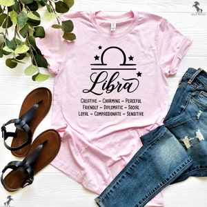 Pu&ograve; includere: Maglietta rosa con un segno zodiacale Bilancia nero e le parole "Creative ~ Charming ~ Peaceful ~ Friendly ~ Diplomatic ~ Social ~ Loyal ~ Compassionate ~ Sensitive".