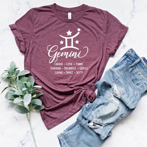 Op de afbeelding: Een maroon t-shirt met een witte afbeelding van het sterrenbeeld Tweelingen en de tekst "Tweelingen" met een lijst van persoonlijkheidstrekken: "Nieuwsgierig - Loyal - Grappig, Grootmoedig - Diplomatiek - Vriendelijk, Liefdevol - Slim - Grappig"