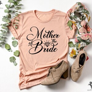 May include: A peach-coloured t-shirt with the text "Mother of the Bride" in black script with a diamond graphic above the word "Bride".