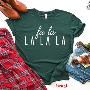 May include: A green t-shirt with white text that reads "fa la LA LA LA". The shirt is laid flat on a white surface with a red and black checked shirt, a pair of brown boots, and a pair of blue jeans.