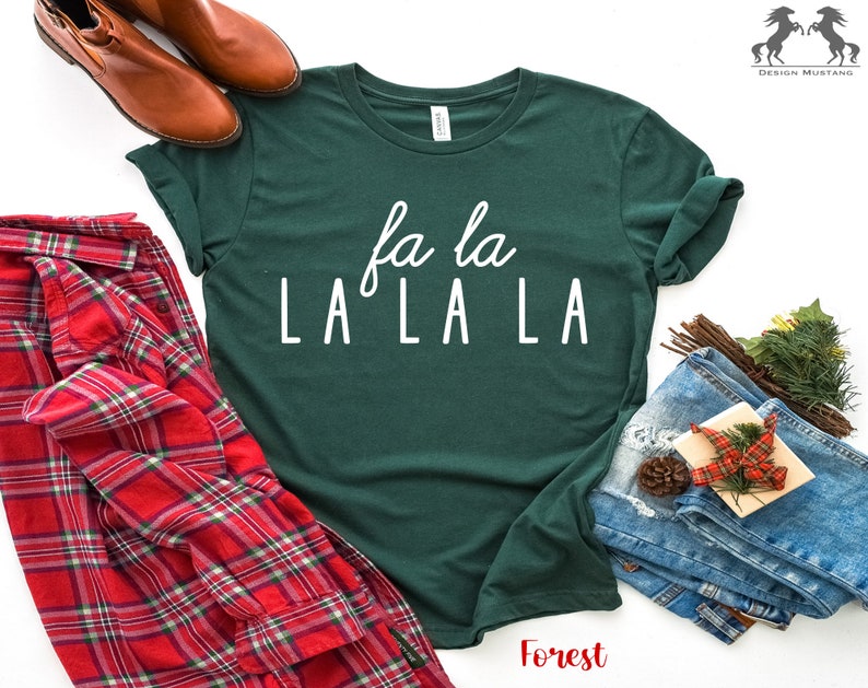 May include: A green t-shirt with white text that reads "fa la LA LA LA". The shirt is laid flat on a white surface with a red and black checked shirt, a pair of brown boots, and a pair of blue jeans.