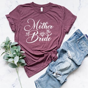 May include: A maroon t-shirt with the text "Mother of the Bride" in white cursive lettering with a diamond graphic. The shirt is laid flat on a white marble surface with a pair of blue jeans.