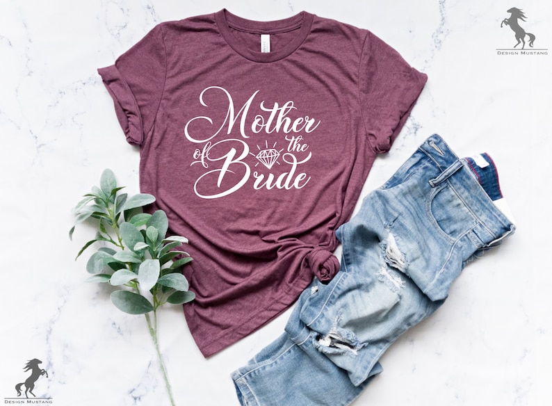 May include: A maroon t-shirt with the text "Mother of the Bride" in white cursive lettering with a diamond graphic. The shirt is laid flat on a white marble surface with a pair of blue jeans.