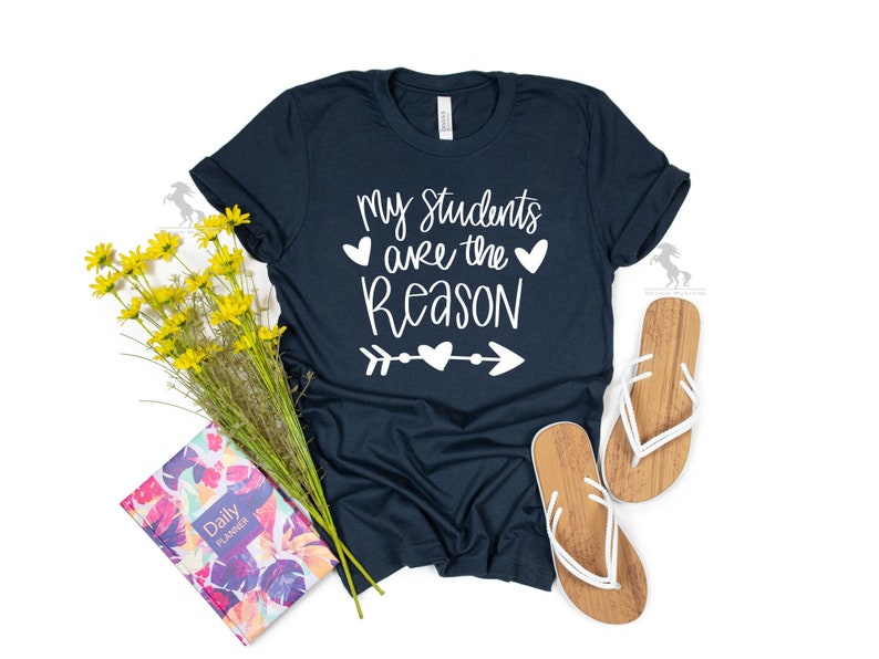 Op de afbeelding: Een marineblauw T-shirt met witte tekst die "Mijn leerlingen zijn de reden" luidt, met een hart- en pijl-afbeelding.