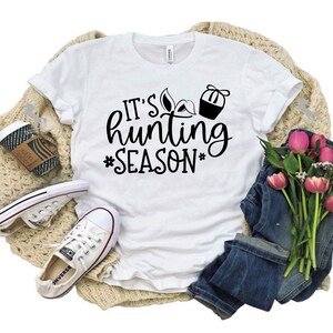K&ouml;nnte beinhalten: Wei&szlig;es T-Shirt mit schwarzem Text, der "It's hunting season" lautet, mit einem Jagdstiefel und einer Truthahn-Grafik. Das Shirt liegt auf einer beigefarbenen Strickdecke mit einem Paar wei&szlig;en Turnschuhen, einer Tasse Kaffee und einem Strau&szlig; rosa Blumen.