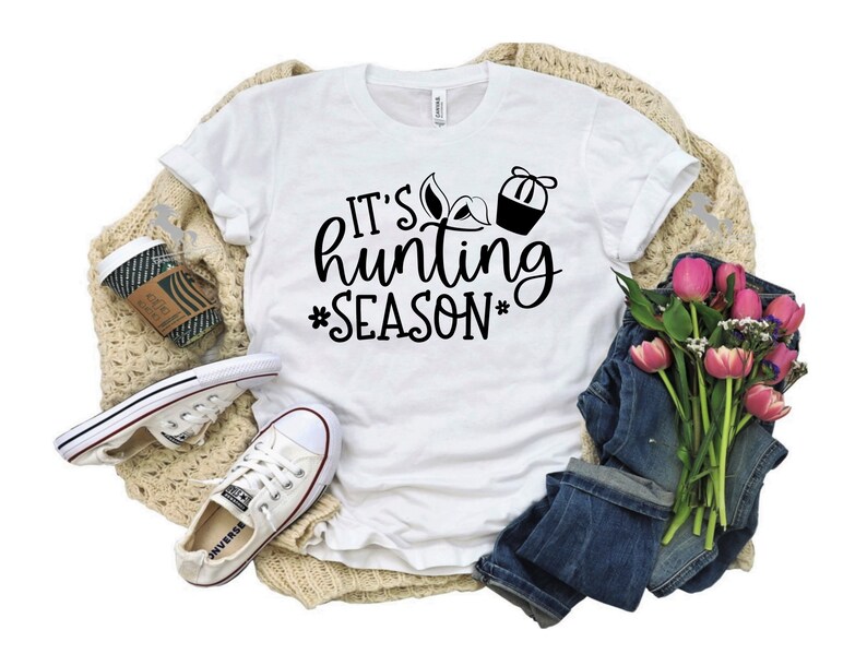 K&ouml;nnte beinhalten: Wei&szlig;es T-Shirt mit schwarzem Text, der "It's hunting season" lautet, mit einem Jagdstiefel und einer Truthahn-Grafik. Das Shirt liegt auf einer beigefarbenen Strickdecke mit einem Paar wei&szlig;en Turnschuhen, einer Tasse Kaffee und einem Strau&szlig; rosa Blumen.