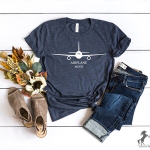 May include: A navy blue t-shirt with a white airplane silhouette and the text "Airplane Mode". The shirt is laid flat on a white wooden surface with a pair of brown sandals and blue jeans.