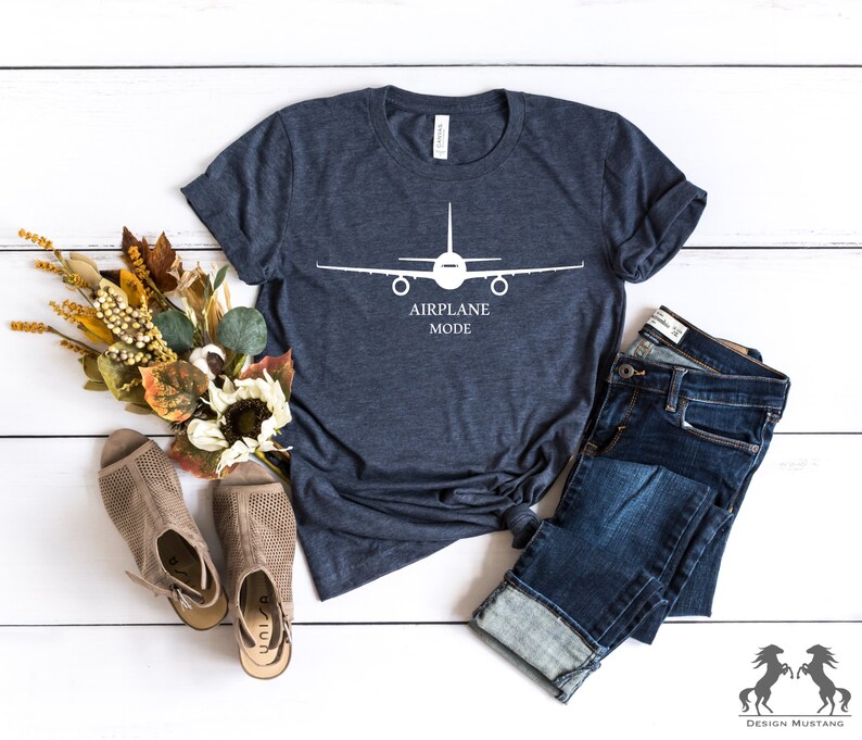 May include: A navy blue t-shirt with a white airplane silhouette and the text "Airplane Mode". The shirt is laid flat on a white wooden surface with a pair of brown sandals and blue jeans.