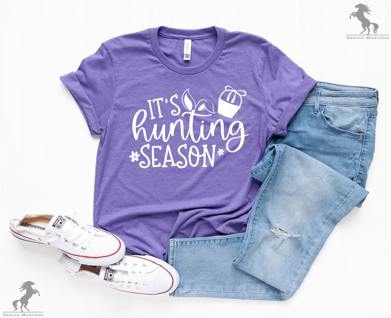 K&ouml;nnte beinhalten: Lilafarbenes T-Shirt mit wei&szlig;em Text, der "It's hunting season" lautet, mit einer kleinen Grafik eines Jagdkorbs und Bl&auml;ttern.