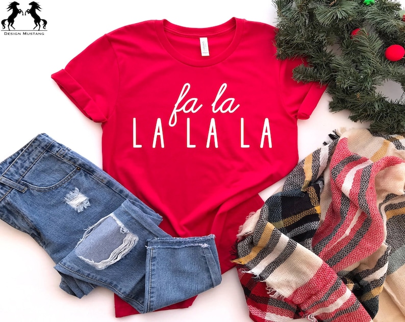 May include: Red t-shirt with white text that says "fa la LA LA LA" on a white background. The shirt is styled with blue jeans and a plaid scarf.