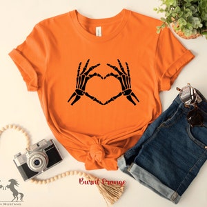 May include: A burnt orange t-shirt with a black skeleton hand design making a heart shape. The shirt is laid flat on a white surface with a camera, wooden beads, and denim shorts.
