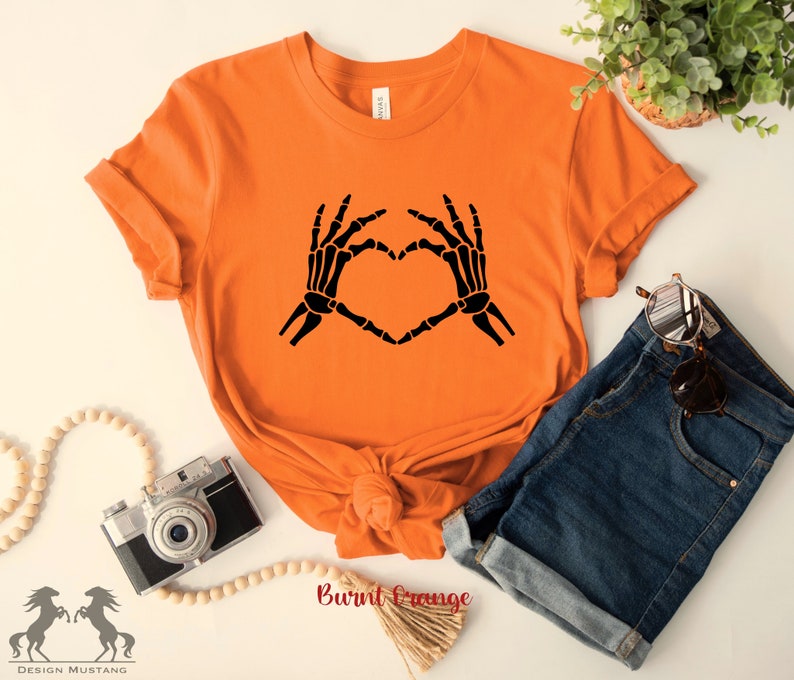 May include: A burnt orange t-shirt with a black skeleton hand design making a heart shape. The shirt is laid flat on a white surface with a camera, wooden beads, and denim shorts.