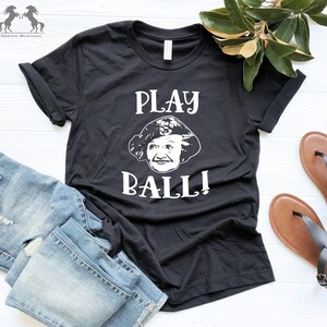 Peut inclure: T-shirt noir avec un graphique blanc d'un visage de femme et le texte "PLAY BALL!"
