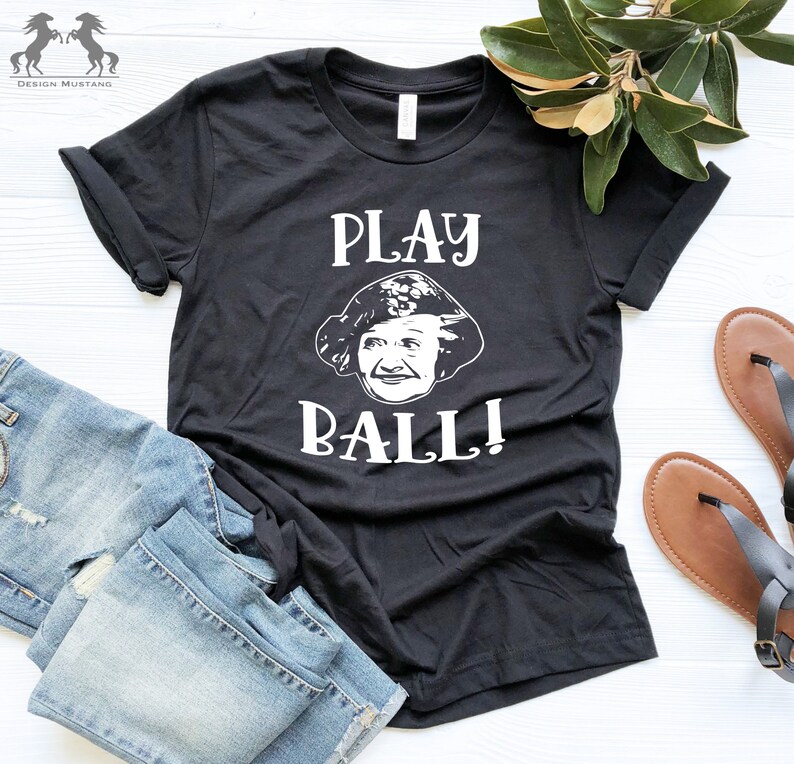 Peut inclure: T-shirt noir avec un graphique blanc d'un visage de femme et le texte "PLAY BALL!"