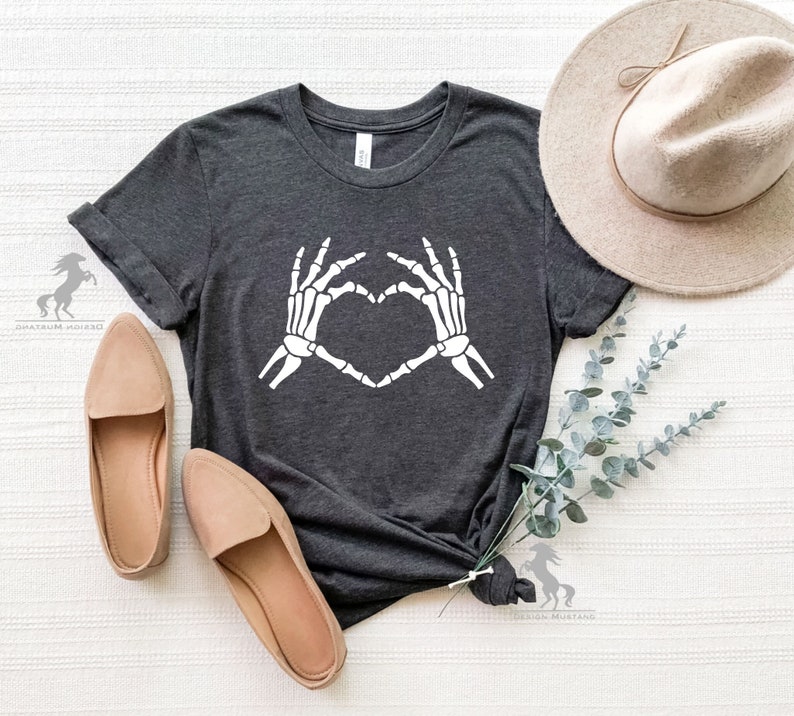 May include: A dark gray t-shirt with a white skeleton hand design making a heart shape. The shirt is laying flat on a white surface with a hat and greenery.