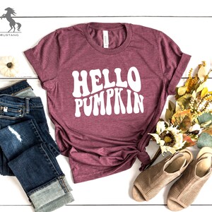 May include: A maroon t-shirt with the text "HELLO PUMPKIN" in white, styled with blue jeans and brown sandals. The shirt has a relaxed fit and a crew neck.