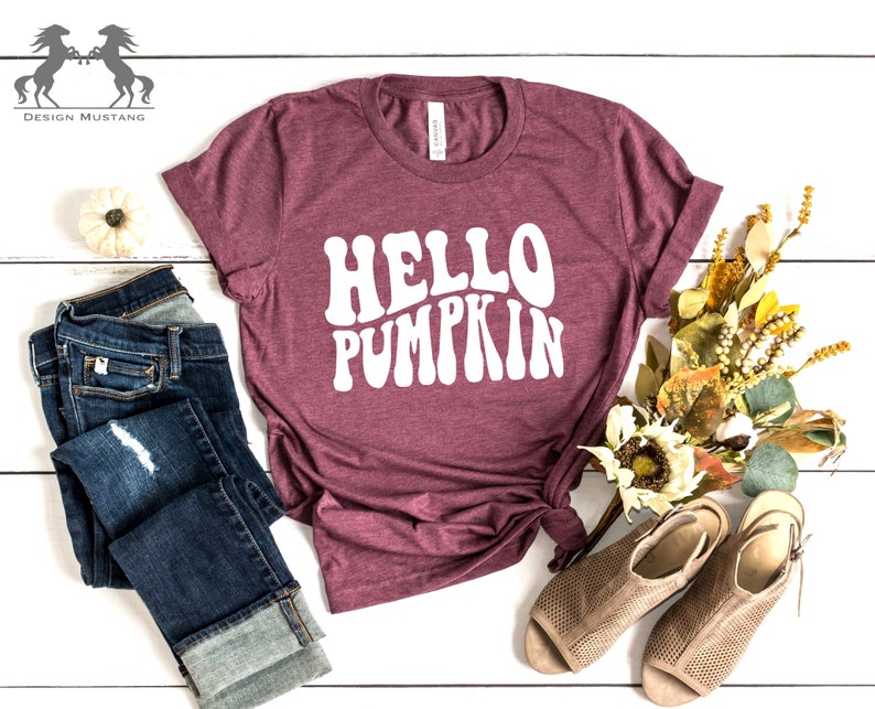 May include: A maroon t-shirt with the text "HELLO PUMPKIN" in white, styled with blue jeans and brown sandals. The shirt has a relaxed fit and a crew neck.