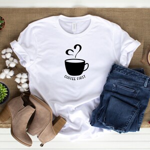 May include: White t-shirt featuring a black graphic of a coffee cup with steam and the text "Coffee First". The shirt is laid out on a brown hessian surface with a pair of brown suede ankle boots, a pair of blue jeans, and a small potted succulent plant.
