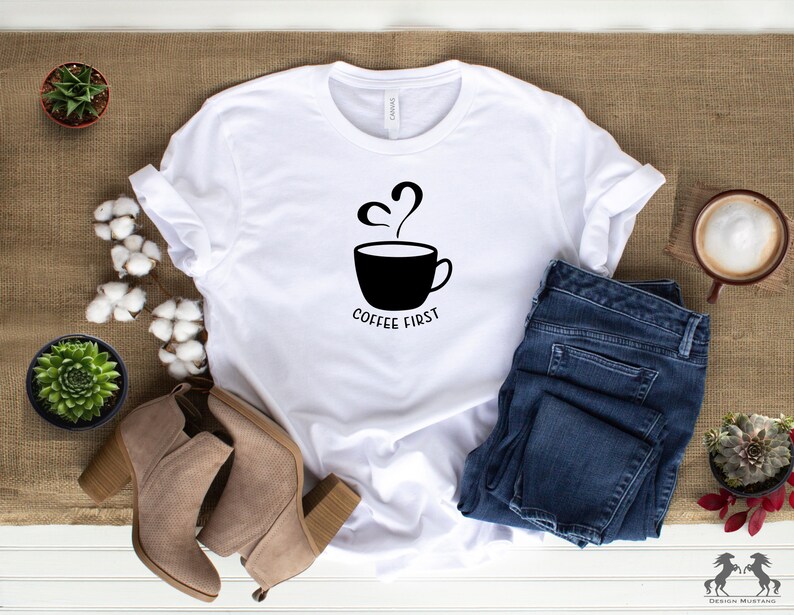 May include: White t-shirt featuring a black graphic of a coffee cup with steam and the text "Coffee First". The shirt is laid out on a brown hessian surface with a pair of brown suede ankle boots, a pair of blue jeans, and a small potted succulent plant.