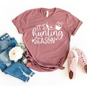 K&ouml;nnte beinhalten: Ein mauvefarbenes T-Shirt mit dem Text "It's hunting season" in wei&szlig;en Buchstaben. Das Shirt liegt flach auf einer wei&szlig;en Oberfl&auml;che mit einem Paar rosa Sneakers und einer blauen Jeans.