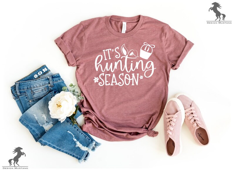 K&ouml;nnte beinhalten: Ein mauvefarbenes T-Shirt mit dem Text "It's hunting season" in wei&szlig;en Buchstaben. Das Shirt liegt flach auf einer wei&szlig;en Oberfl&auml;che mit einem Paar rosa Sneakers und einer blauen Jeans.