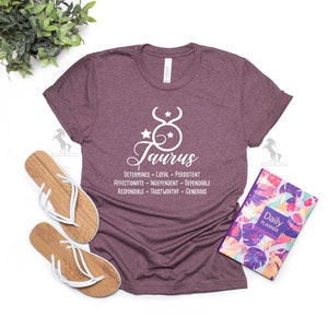 May include: A maroon t-shirt with a white graphic of the Taurus zodiac sign and the word "Taurus" in cursive. The graphic also includes a list of Taurus personality traits: "Determined - Loyal - Persistent - Affectionate - Independent - Dependable - Responsible - Trustworthy - Generous."