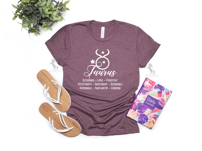 May include: A maroon t-shirt with a white graphic of the Taurus zodiac sign and the word "Taurus" in cursive. The graphic also includes a list of Taurus personality traits: "Determined - Loyal - Persistent - Affectionate - Independent - Dependable - Responsible - Trustworthy - Generous."