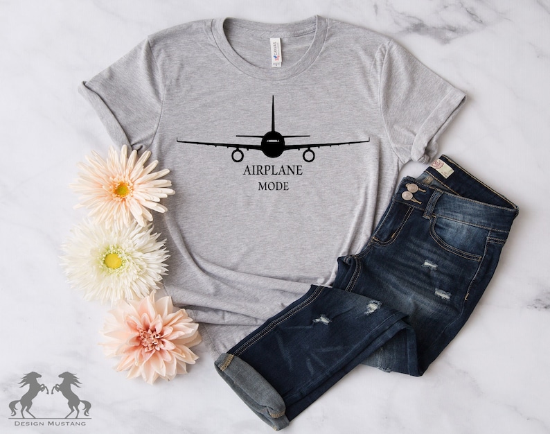 May include: A gray t-shirt with a black airplane silhouette and the text "AIRPLANE MODE" printed on the front. The shirt is laid out on a white surface with a pair of blue jeans and flowers.