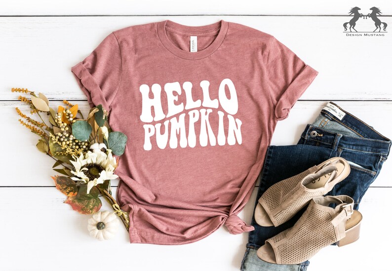 May include: A mauve t-shirt with the text "HELLO PUMPKIN" in white, retro-style lettering. The shirt is laid flat on a white wooden surface with a bouquet of fall foliage and a white pumpkin.