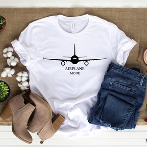 May include: White t-shirt with a black airplane silhouette and the text "AIRPLANE MODE". The shirt is laid out on a brown burlap surface with blue jeans, brown boots, and a cup of coffee.
