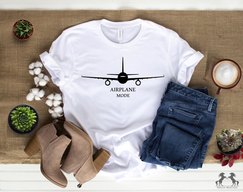 May include: White t-shirt with a black airplane silhouette and the text "AIRPLANE MODE". The shirt is laid out on a brown burlap surface with blue jeans, brown boots, and a cup of coffee.