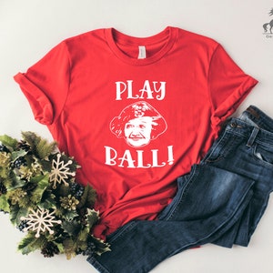 Peut inclure: T-shirt rouge avec un graphique blanc d'un visage de femme et le texte "PLAY BALL!"