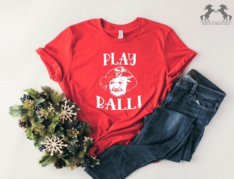 Peut inclure: T-shirt rouge avec un graphique blanc d'un visage de femme et le texte "PLAY BALL!"