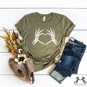 May include: A green t-shirt with a white skeleton hand design making a heart shape. The shirt is laid out on a white wooden surface with a pair of brown sandals, a bouquet of dried flowers, and a pair of blue jeans.