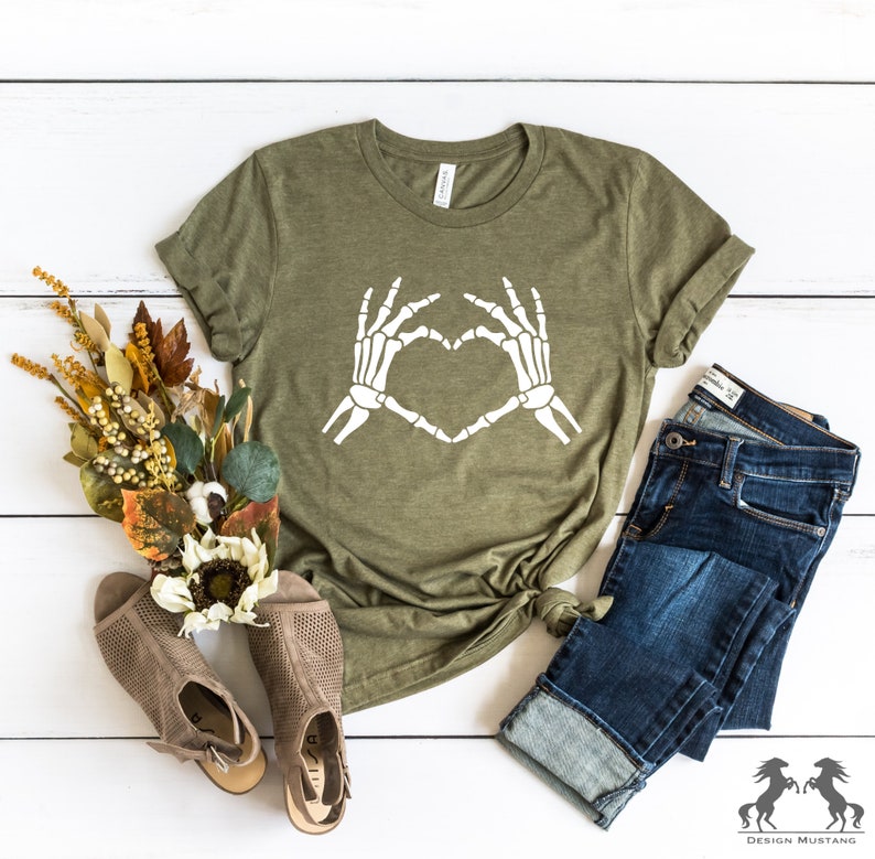 May include: A green t-shirt with a white skeleton hand design making a heart shape. The shirt is laid out on a white wooden surface with a pair of brown sandals, a bouquet of dried flowers, and a pair of blue jeans.