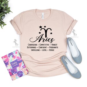 Może przedstawiać: Jasnor&oacute;żowy T-shirt z czarną grafiką przedstawiającą znak zodiaku Baran ze gwiazdami i słowem "Baran" pisanym kursywą. Tekst pod grafiką brzmi: "Odważny - Konkurencyjny - Szczery - Zdeterminowany - Pewny siebie - Pasjonat - Nieugięty - Lojalny - Bohaterski."