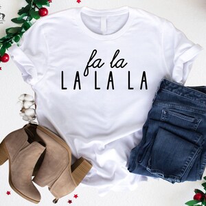 May include: White t-shirt with the text "fa la LA LA LA" in black lettering. The shirt is styled with blue jeans and brown ankle boots.