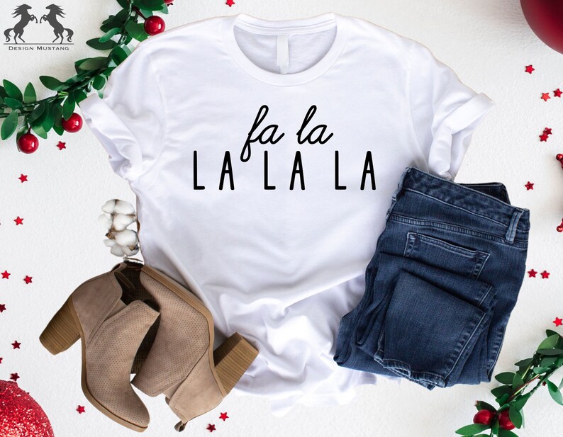 May include: White t-shirt with the text "fa la LA LA LA" in black lettering. The shirt is styled with blue jeans and brown ankle boots.