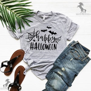 May include: A gray t-shirt with the text "happy HALLOWEEN" in black lettering. The design includes a spider web with a spider, and three bats. The shirt is laid out on a white surface with a pair of brown sandals and blue jeans.