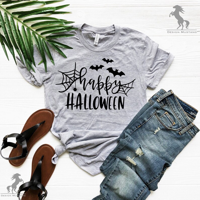 May include: A gray t-shirt with the text "happy HALLOWEEN" in black lettering. The design includes a spider web with a spider, and three bats. The shirt is laid out on a white surface with a pair of brown sandals and blue jeans.