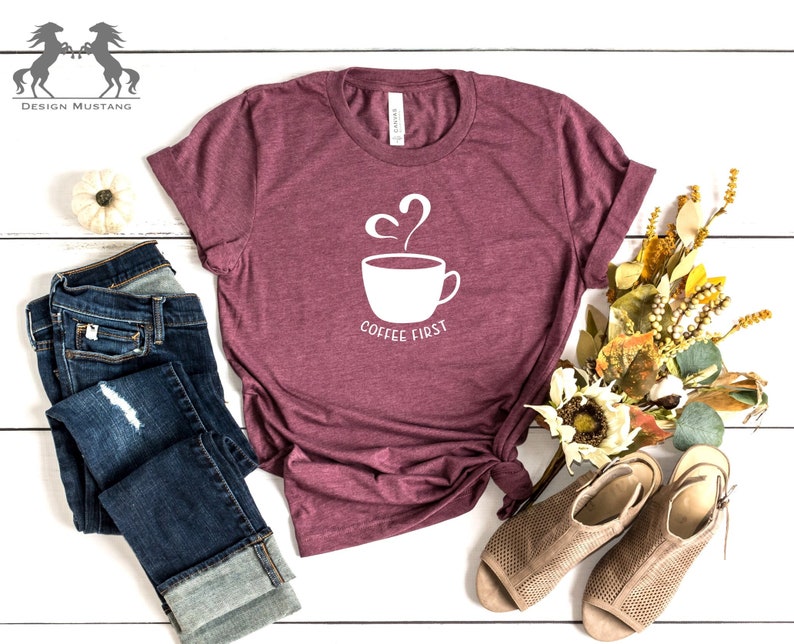 May include: A maroon t-shirt featuring a white graphic of a coffee cup with steam rising from it and the text "Coffee First". The shirt is laid out on a white wooden surface with a pair of blue jeans, a small white pumpkin, and a bouquet of dried flowers.