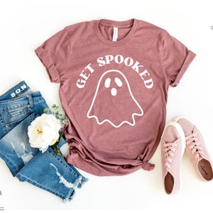 K&ouml;nnte beinhalten: Ein mauvefarbenes T-Shirt mit einer wei&szlig;en Geistergrafik und dem Text "GET SPOOKED". Das Shirt liegt auf einer wei&szlig;en Oberfl&auml;che mit einem Paar rosa Sneakers und einer blauen Jeans.